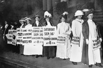 A black and white photo of women in dresses and feathered hats. They hold signs advertising an upcoming rally for women's right to vote.