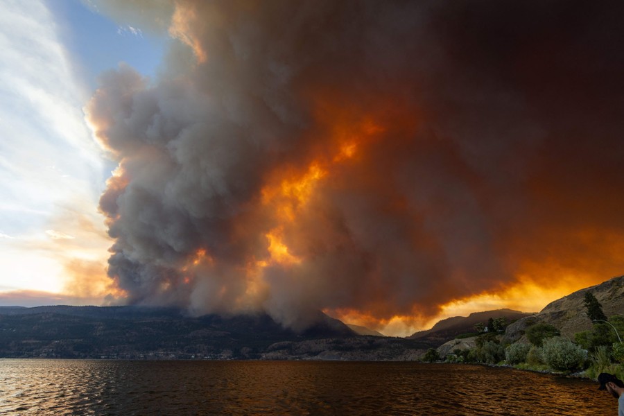 Huge clouds of smoke rise above a forest fire near a lake.