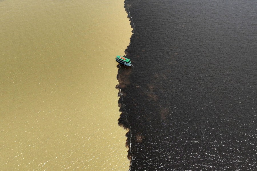 A drone view of a boat passing through the confluence of two rivers, with the left side of the photo dominated by muddy, cream-colored water, and the right side, much darker water.