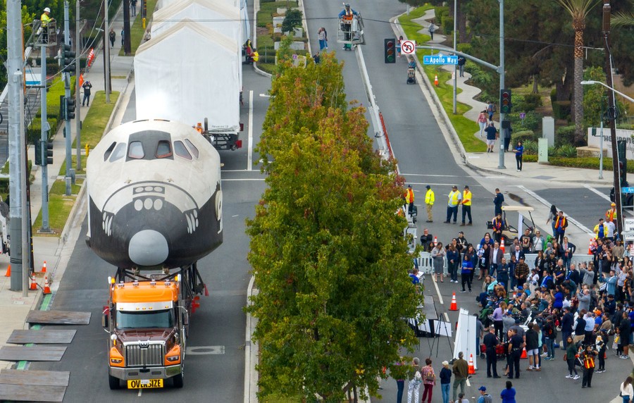 A crowd watches as movers transport pieces of a full-scale mock-up of a space shuttle.