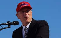 Andy Biggs stands before a microphone wearing a "Trump: Make America Great Again" hat.
