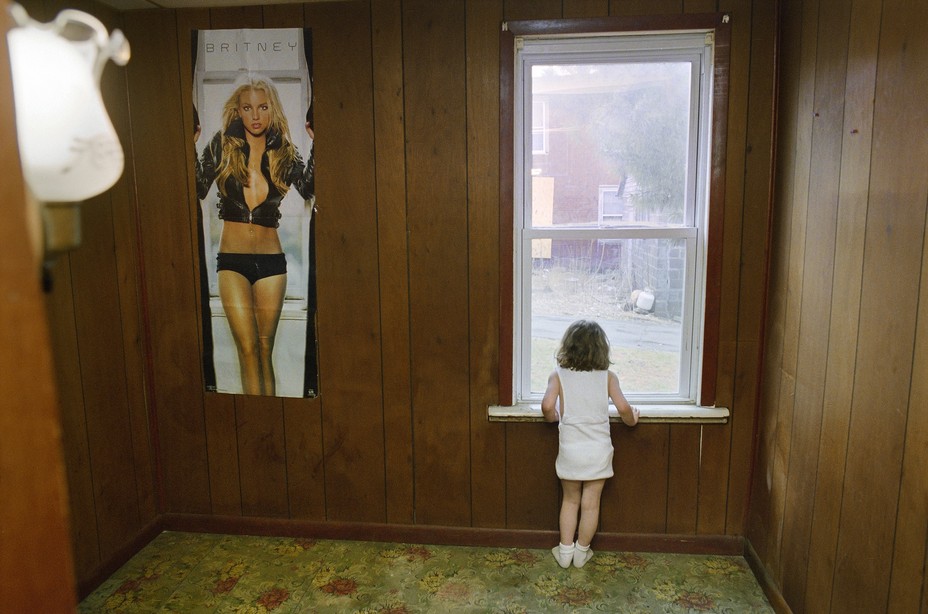 a young girl in an empty room with a Britney Spears poster looks out the window.