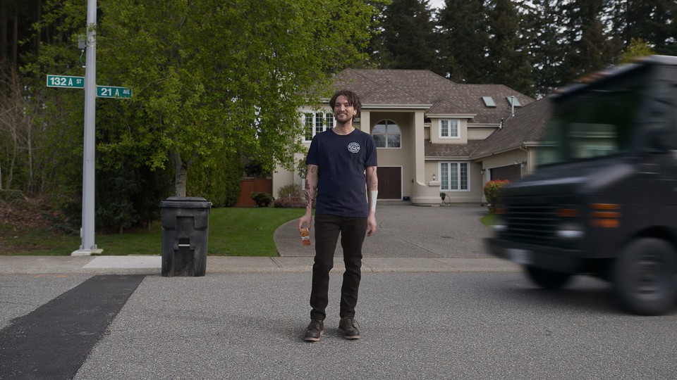 A color film still from "Final Destination Bloodlines" showing a man standing in the middle of a suburban street, looking at the camera, while a big truck approaches behind him from the right.