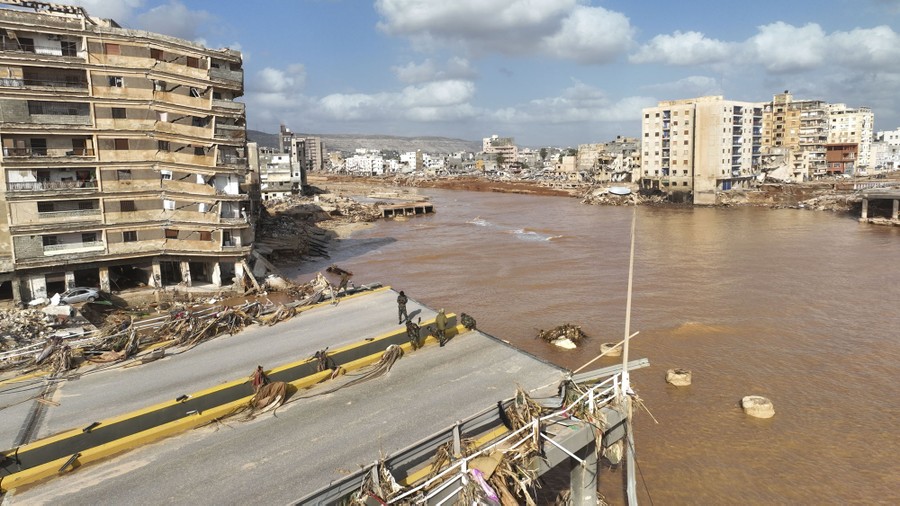 Several people stand on what remains of a road bridge that has been washed away by a flood, beside many damaged buildings.