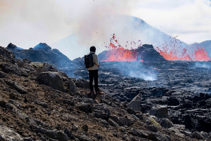A person stands on a hillside looking toward lava erupting nearby.