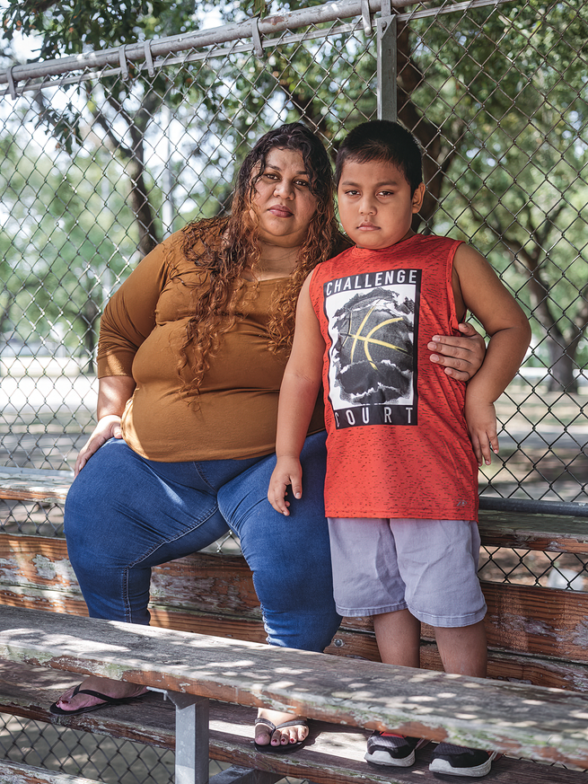 Photo of a woman sitting on wooden bleachers in front of a chain link fence, with her arm around a small boy standing next to her