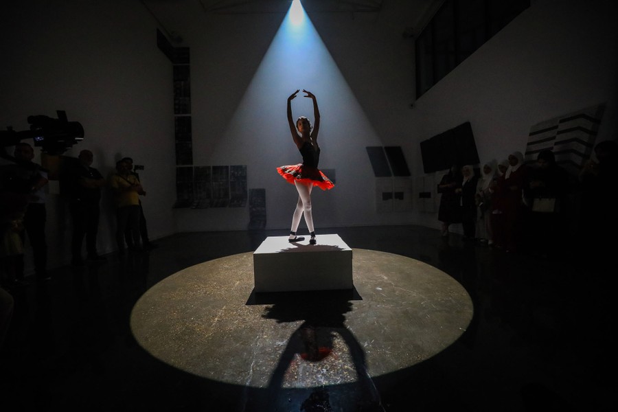 A ballerina performs on a tiny stage in a performance space, lit by a single light above.