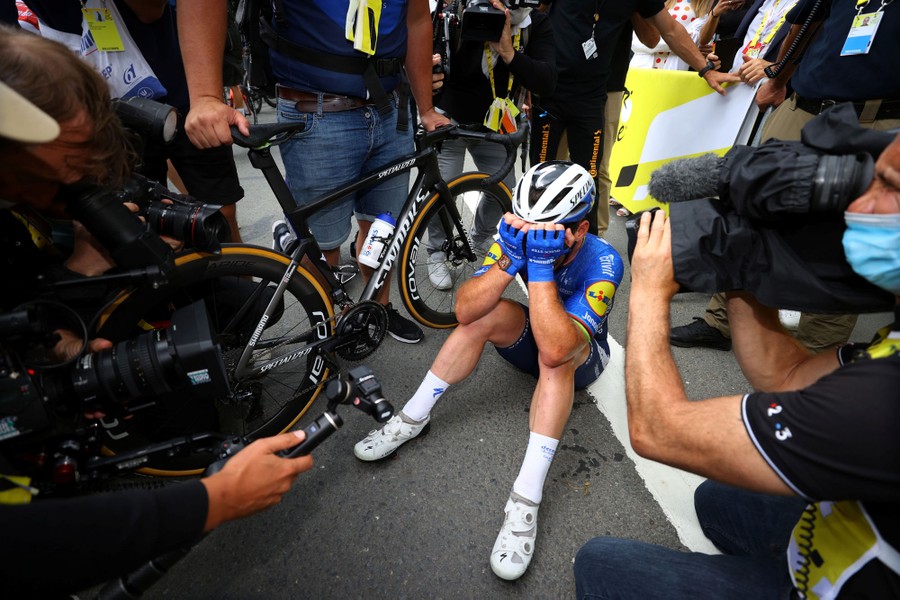 A cyclist sits on the pavement, hands over his face, surrounded by onlookers and photographers.