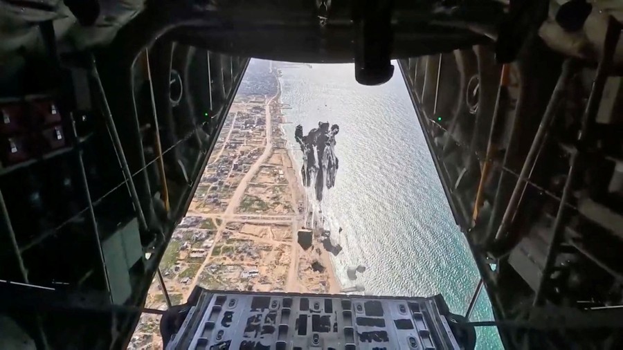 A view out the open rear door of a military aircraft as aid packages attached to parachutes are dropped over a beach, seen below