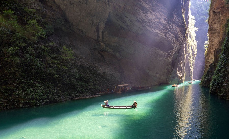 People ride small boats on clear water in a tall and narrow canyon.