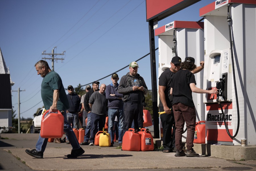 About a dozen people stand in line at a gas-station pump, with red plastic gas cans.