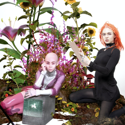 Grimes, in an orange wig, sits amid a field of flowers.