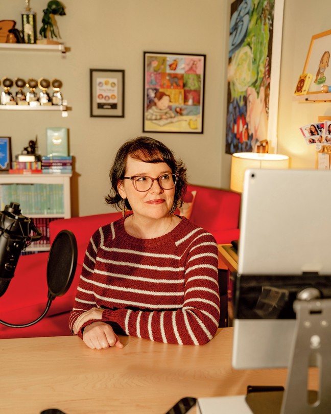 a portrait of the cartoonist and author Raina Telgemeier, wearing a red striped sweater and sitting in her home, in front of a bookshelf of her work.
