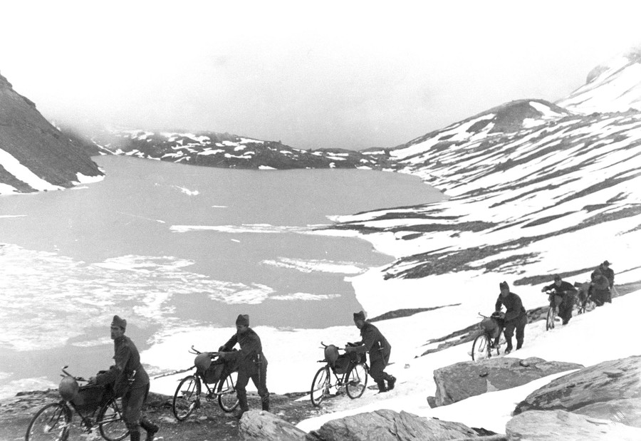 A half-dozen soldiers walk beside their bicycles through a partially snow-covered mountain pass.