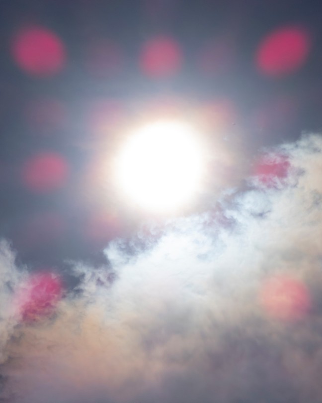 An photo of wispy clouds with a blurry sun, and pink dots across the image