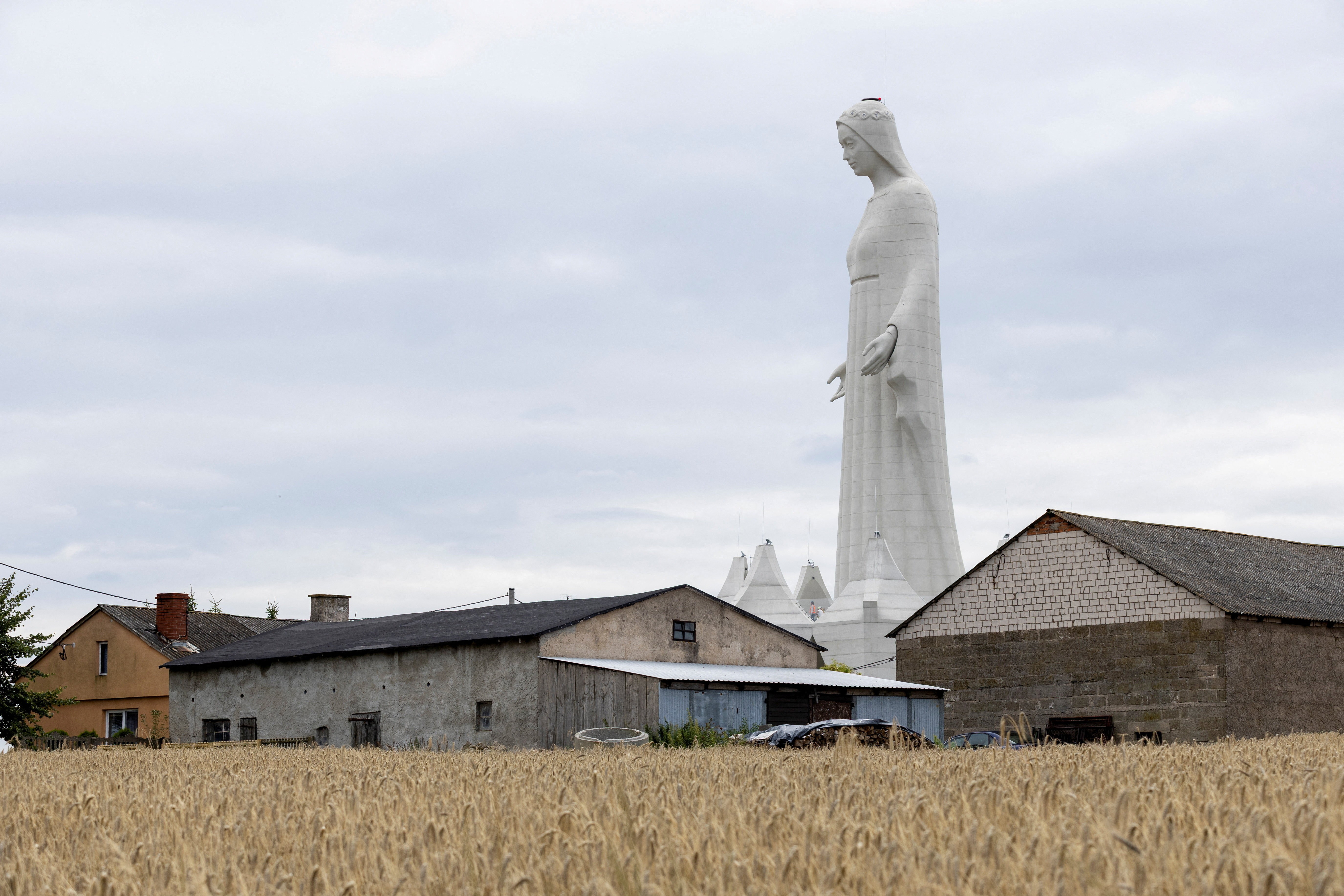 A very tall statue of the Virgin Mary stands near several farm buildings.