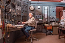 photo of two uniformed men sitting in control center with huge analog machinery bank in background