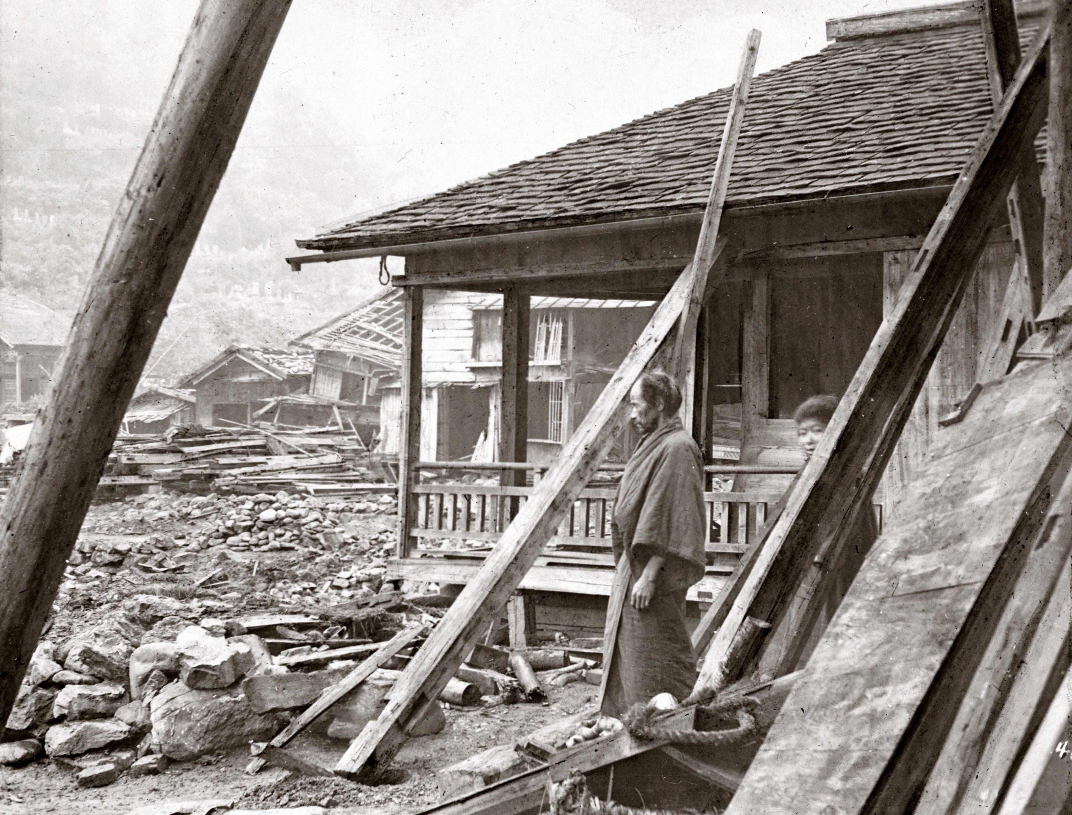 Two people stand outside a damaged house, looking at debris from other earthquake-damaged houses.