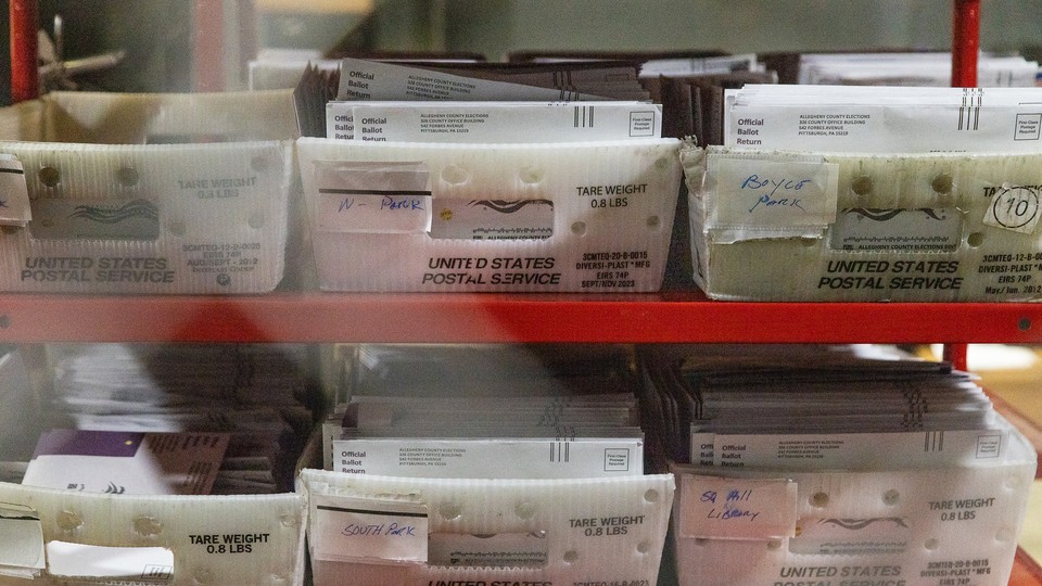 Photograph of mail-in ballots in plastic United States Postal Service bins on a red shelf