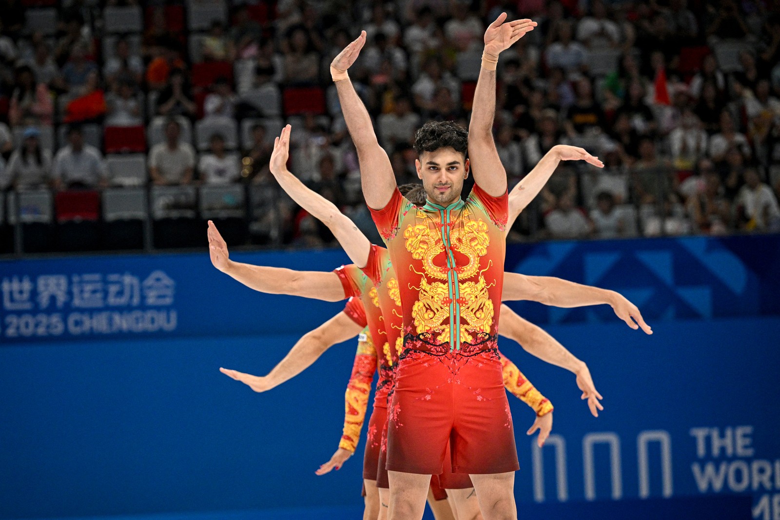 A group of five gymnasts poses during a performance, lined up back-to-front, each with their arms outstretched in varying orientations.