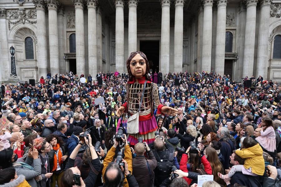 An 11-foot-tall puppet of a young girl stand surrounded by a crowd looking at it in front of a cathedral.