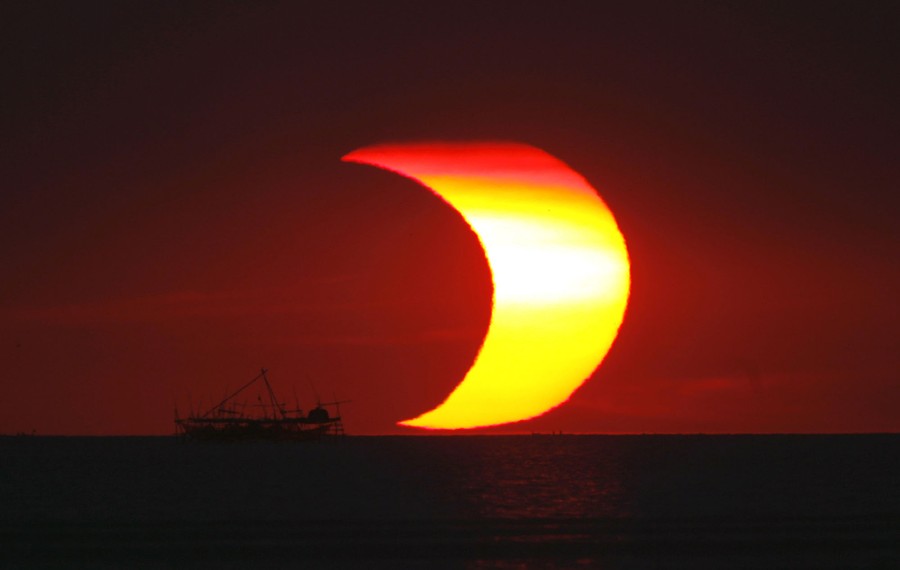 The partly-eclipsed sun, shaped like a crescent, sets over the ocean, seen near a fishing boat.
