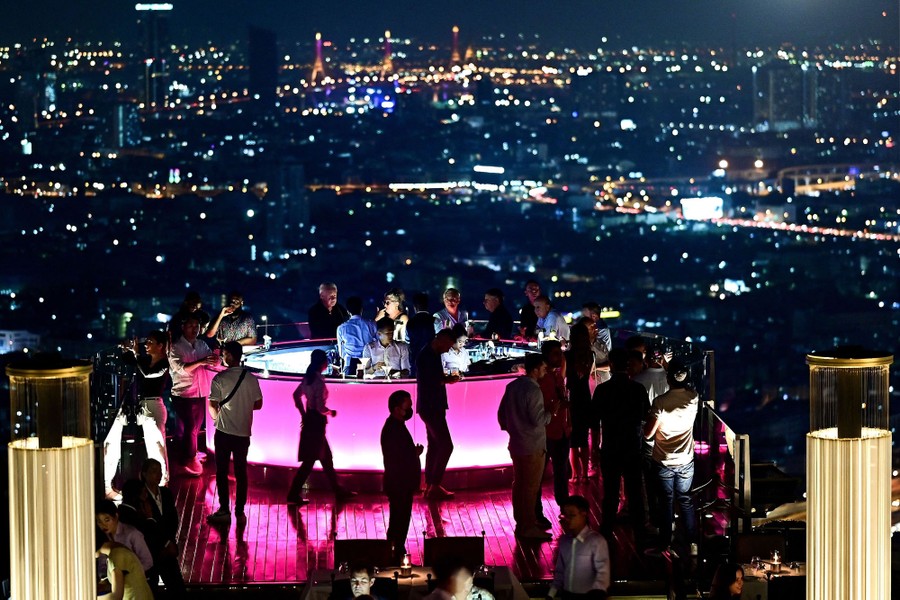 People stand at a rooftop bar, with city lights visible in the background.