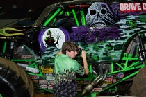 A young man stands beside a monster truck that features a skull on its side.