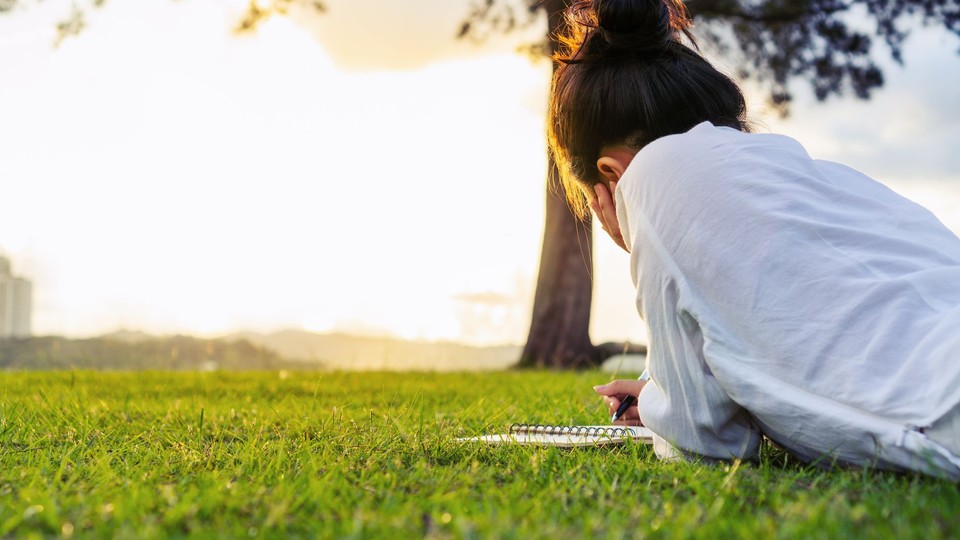 rear view of woman with black hair in a bun lying on her stomach on green grass, writing in a noteboo