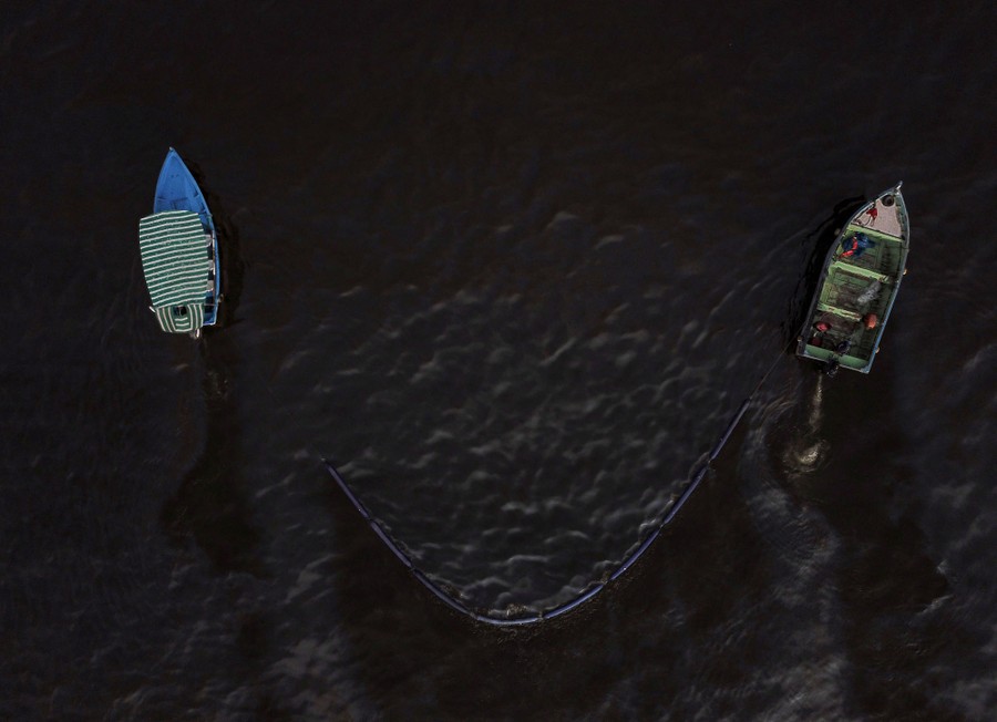 An aerial view of two small boats towing a boom, collecting floating oil.