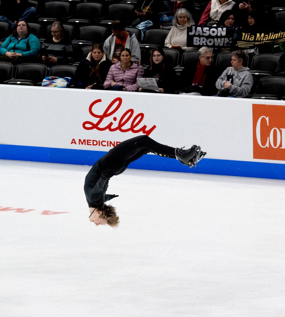 photo of ice rink with skater dressed in black in mid-flip upside down with legs and arms extended