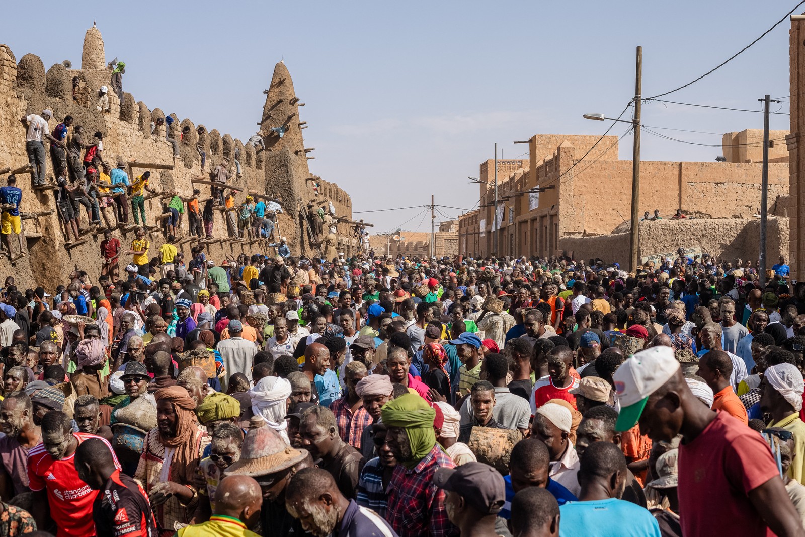 A large crowd of people gather around an ancient mosque, built of mud and brick, repairing and replastering it.