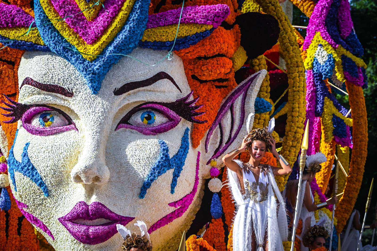 People ride on a parade float shaped like a person's face covered in colorful flowers.