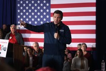A photo of Ron DeSantis standing in front of a small, seated crowd and an American flag