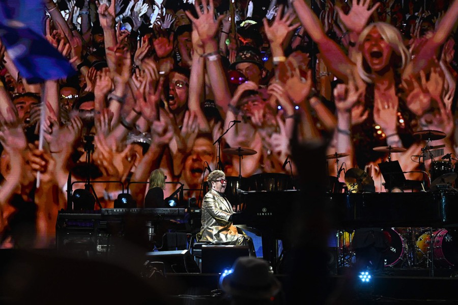 A cheering crowd on a giant video display behind a performer who sings and plays piano on a stage.