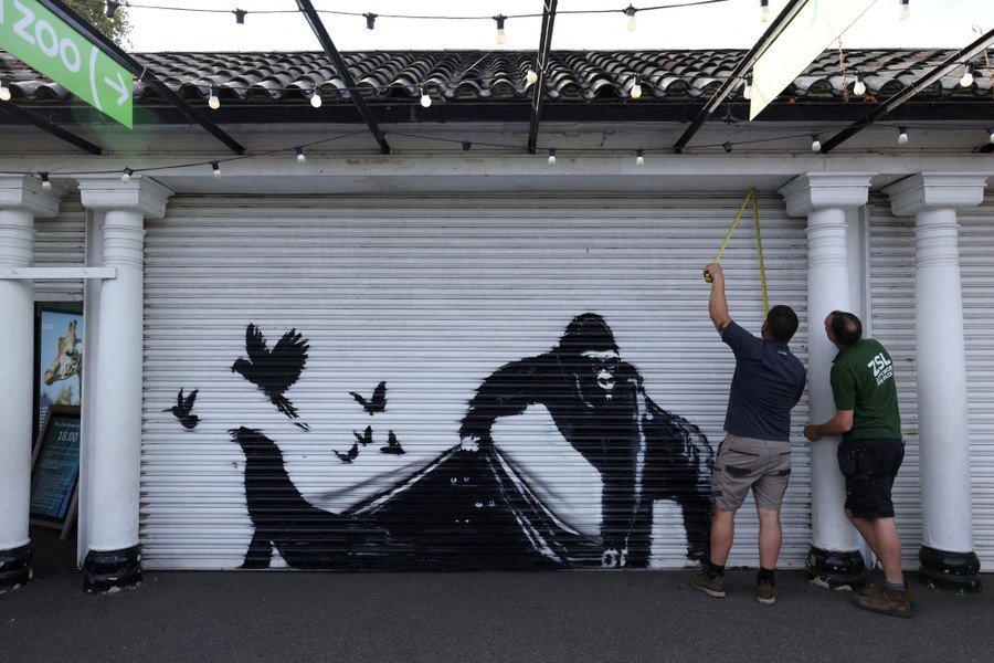 Two people use a measuring tape beside a stencil painting of a gorilla on a rolling door.