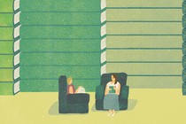 Two women sit in comfortable armchairs, in front of a wall of giant books