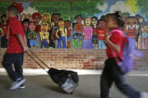 Two black children walk with backpacks past a mural depicting a diverse array of children.