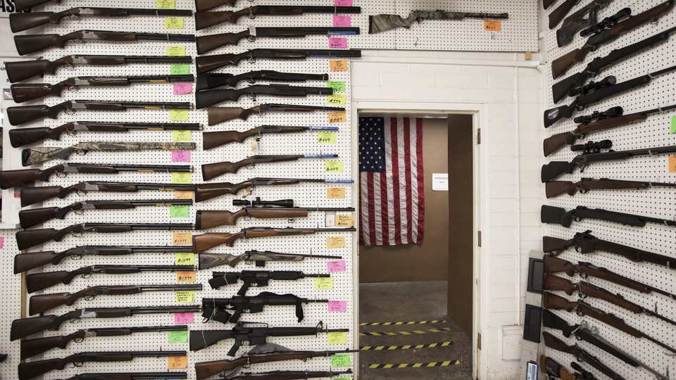 A photograph taken inside a gun shop, in which columns of rifles hang on white pegboard walls. An American flag is visible through an open doorawy.