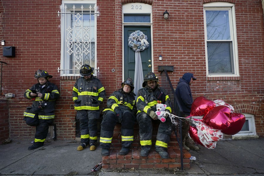 Several firefighters gather outside a house, heads down, as one holds a stuffed bear and several heart balloons.
