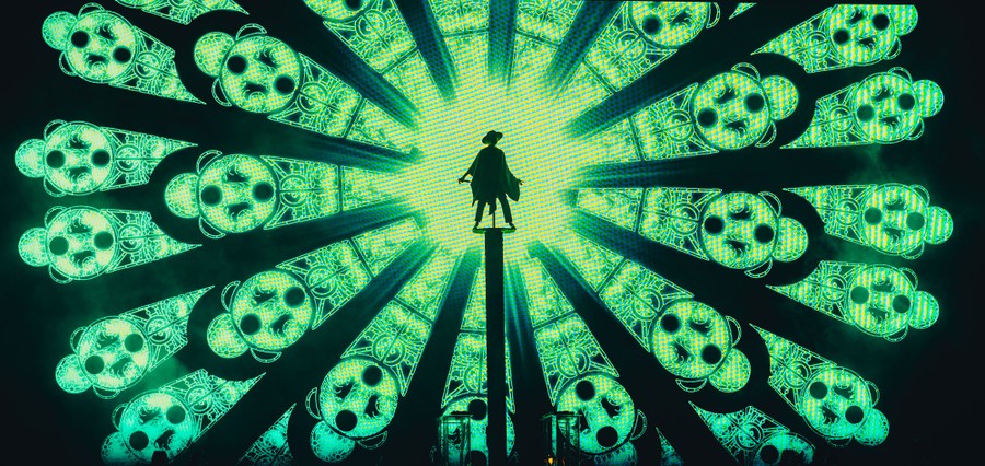 A performer stands on a platform in front of a large video screen displaying a bright radial pattern.