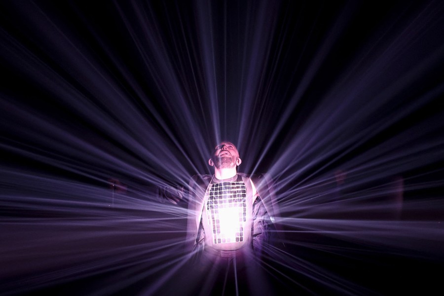 A performer stands on a dark stage wearing a mirrored shirt, reflecting light.