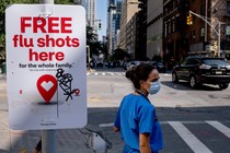 Photo of a woman on a city sidewalk wearing medical scrubs and a mask, next to a sign promoting free flu shots