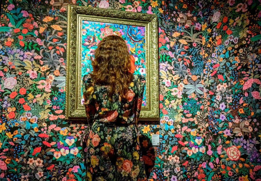 A woman in a floral print outfit stands in front of a wall and painting covered in floral print.