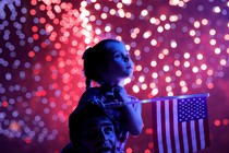 A young girl sits on the shoulders of an adult, holding an American flag, with fireworks in the background.
