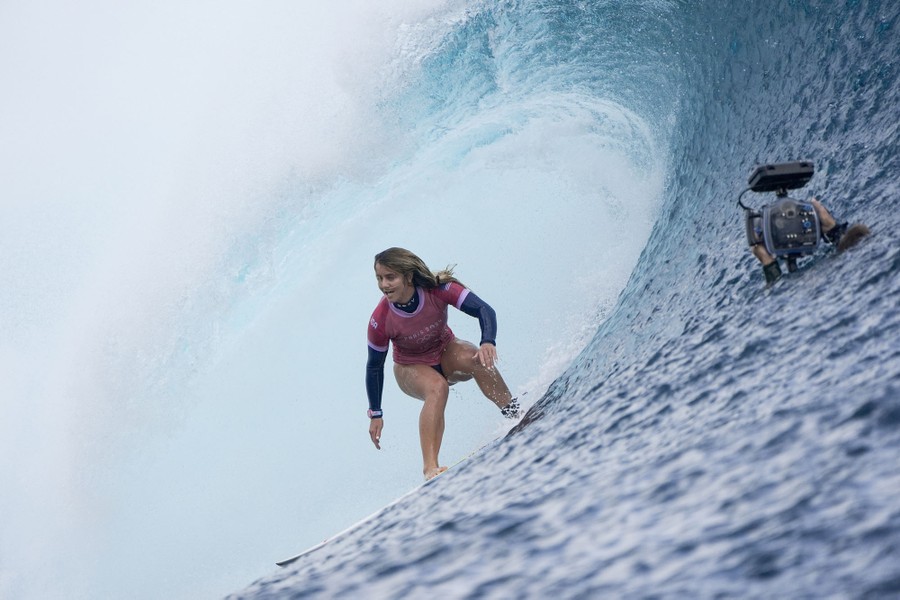 A surfer rides in the curl of a large wave, with a mostly submerged photographer holding up a waterproof camera nearby.
