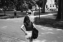 A black-and-white photo of a young woman, seen from behind, wearing a backpack and walking on a campus