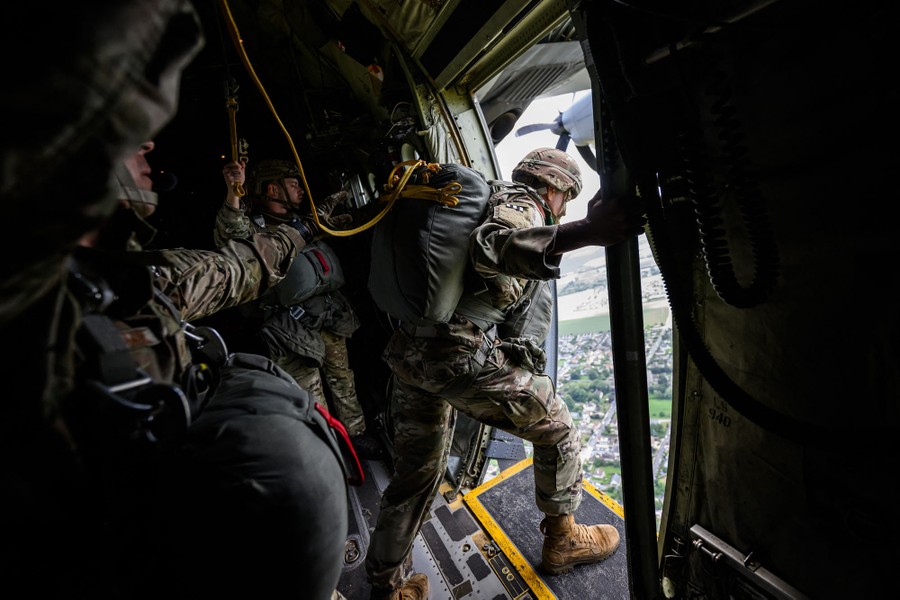 Several soldiers wearing parachutes line up in an airborne aircraft, beside an open door.
