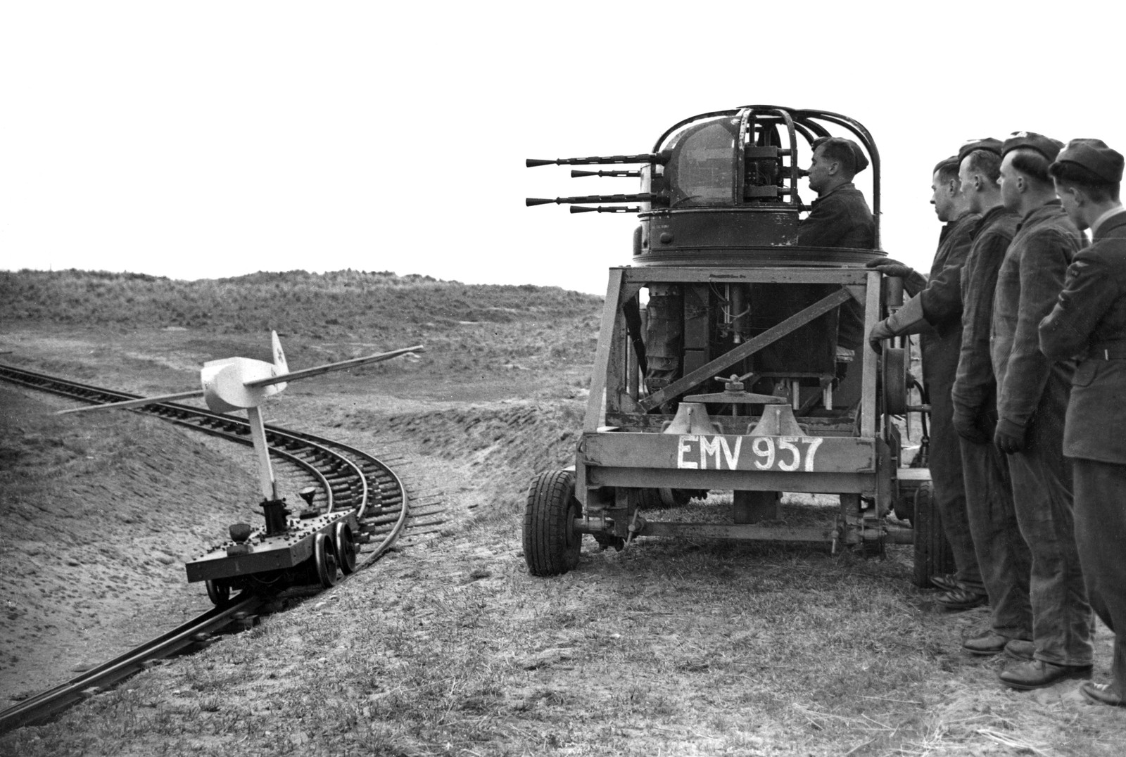 A group of soldiers stands near one soldier who is sitting in a mounted machine gun apparatus, as a model plane mounted to a track passes by.