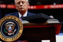 Photograph of Donald Trump behind a desk with the presidential seal and executive orders atop the desk